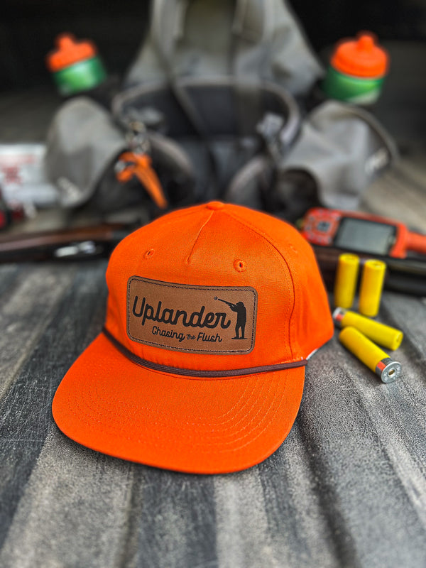 Orange hunting cap with a brand logo on a gray surface with hunting equipment in the background.