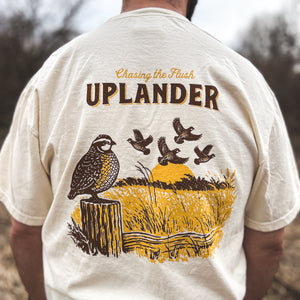 Back view of a man wearing an off-white t-shirt featuring a brown and gold graphic of a Bobwhite Quail perched on a fence post and a covey of quail flushing in the distance, with text 'Chasing the Flush, Uplander', worn outdoors.