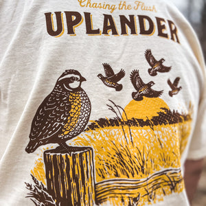 Close-up back view of a man wearing an off-white t-shirt featuring a brown and gold graphic of a Bobwhite Quail perched on a fence post and a covey of quail flushing in the distance, with text 'Chasing the Flush, Uplander', worn outdoors.