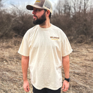 Front view of a man wearing an off-white t-shirt with text 'Uplander, Chasing the Flush' on the upper left chest, worn outdoors.