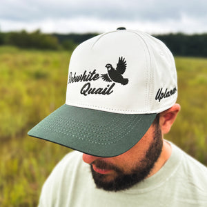Man wearing an ivory and dark green two-tone snapback hat with an embroidered flying bobwhite quail and 'Bobwhite Quail' script on the front, 'Uplander' embroidered on the side, worn outdoors.