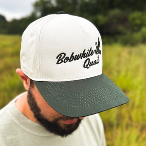Right-side view of a man wearing an ivory and dark green two-tone snapback hat with an embroidered flying bobwhite quail and 'Bobwhite Quail' script on the front, worn outdoors.