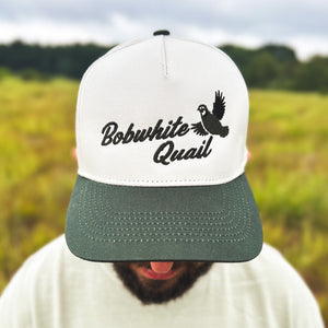Close-up view of a man wearing an ivory and dark green two-tone snapback hat with an embroidered flying bobwhite quail and 'Bobwhite Quail' script on the front, worn outdoors.