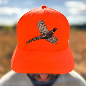 Close-up view of a man wearing a blaze orange snapback hat featuring an embroidered flying rooster pheasant on the front, worn outdoors.