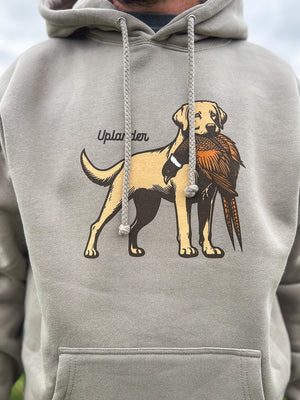 Close-up view of a man wearing a grey hoodie featuring a graphic of a yellow Labrador Retriever holding a ring-necked pheasant in its mouth with Uplander branding, worn outdoors.