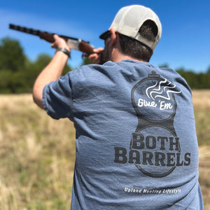 Back view of a man aiming a shotgun and wearing a blue t-shirt featuring a graphic of a smoking shotgun breech with 'Give 'Em Both Barrels' text and Uplander branding, worn outdoors.