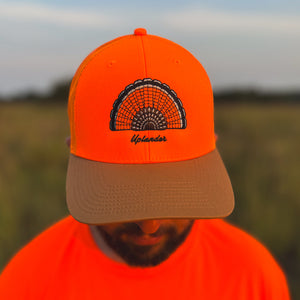 Man wearing a blaze orange snapback hat with a mesh back and a light brown bill featuring an embroidered ruffed grouse tail fan with the script 'Uplander' below, worn outdoors.