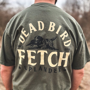 Back view of a man wearing an olive green t-shirt featuring a bird dog retrieving a ring-necked pheasant with text 'Dead Bird Fetch' and Uplander branding, worn outdoors.