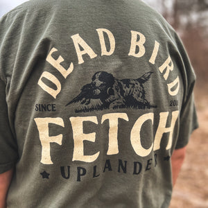 Close-up back view of a man wearing an olive green t-shirt featuring a bird dog retrieving a ring-necked pheasant with text 'Dead Bird Fetch' and Uplander branding, worn outdoors.