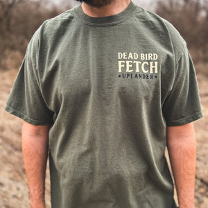Front view of a man wearing an olive green t-shirt with text 'Dead Bird Fetch, Uplander' on the upper left chest, worn outdoors.
