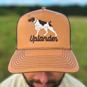 Close-up view of a man wearing a caramel and black trucker hat with an embroidered liver and white German Shorthaired Pointer and 'Uplander' script in brown, worn outdoors.