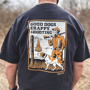 Back view of a man wearing a black t-shirt featuring a vintage-style graphic of a skeleton hunter and bird dog with text 'Good Dogs Crappy Shooting' and Uplander branding, worn outdoors.