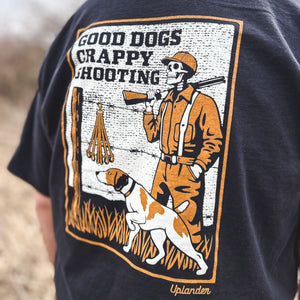 Close-up back view of a man wearing a black t-shirt featuring a vintage-style graphic of a skeleton hunter and bird dog with text 'Good Dogs Crappy Shooting' and Uplander branding, worn outdoors.