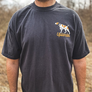Front view of a man wearing a black t-shirt featuring a bird dog graphic with text 'Uplander' on the upper left chest, worn outdoors.