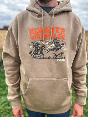 Man wearing a tan hoodie featuring a graphic of a man riding a horse lassoing a pheasant with his bird dog, with text 'Rooster Wrangler, Uplander', worn outdoors.