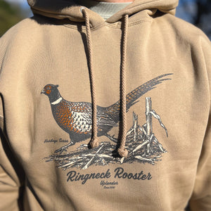 Man wearing a tan heavyweight hoodie featuring a vintage-style illustration of a rooster pheasant in a cornfield, with 'Ringneck Rooster,' 'Uplander,' and 'Heritage Series' script, worn outdoors.