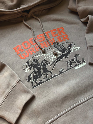 Flat lay of a tan hoodie featuring a graphic of a man riding a horse lassoing a pheasant with his bird dog, with text 'Rooster Wrangler, Uplander'.