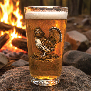 The King Heritage Series 001 pint glass filled with beer, featuring a ruffed grouse illustration, resting on rocks beside a glowing campfire.
