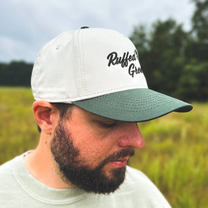 Right-side view of a man wearing an ivory and dark green two-tone snapback hat with an embroidered flying ruffed grouse and 'Ruffed Grouse' script on the front, worn outdoors.