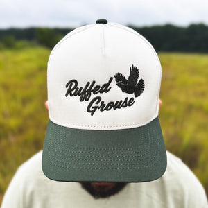 Close-up view of a man wearing an ivory and dark green two-tone snapback hat with an embroidered flying ruffed grouse and 'Ruffed Grouse' script on the front, worn outdoors.
