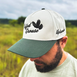 Man wearing an ivory and dark green two-tone snapback hat with an embroidered flying ruffed grouse and 'Ruffed Grouse' script on the front, 'Uplander' embroidered on the side, worn outdoors.