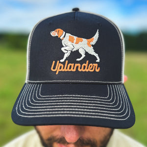 Close-up view of a man wearing a black and cream trucker hat with an embroidered orange and white English Setter and 'Uplander' script in orange, worn outdoors.