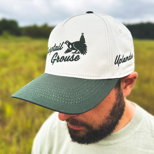 Man wearing an ivory and dark green two-tone snapback hat with an embroidered sharptail grouse and 'Sharptail Grouse' script on the front, 'Uplander' embroidered on the side, worn outdoors.