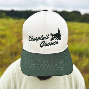 Close-up view of a man wearing an ivory and dark green two-tone snapback hat with an embroidered sharptail grouse and 'Sharptail Grouse' script on the front, worn outdoors.