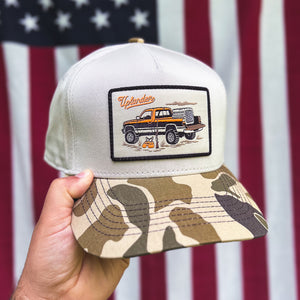 Hand holding a cream-colored trucker hat with a camouflage bill and an embroidered patch featuring a vintage pickup truck and upland hunting gear in front of a blurred American flag.