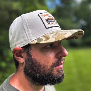 Side view of a man wearing a cream-colored trucker hat with a camouflage bill and an embroidered patch featuring a vintage pickup truck and upland hunting gear, worn outdoors.