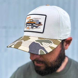 Man wearing a cream-colored trucker hat with a camouflage bill and an embroidered patch featuring a vintage pickup truck and upland hunting gear.