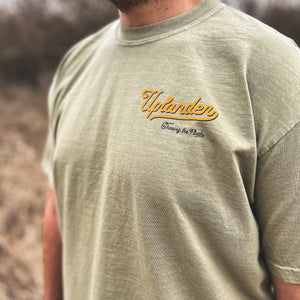 Front chest detail of a khaki t-shirt with text 'Uplander, Chasing the Flush' on the upper left chest.
