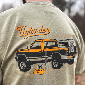 Close-up back view of a man wearing a khaki t-shirt featuring a vintage pickup truck with a dog kennel, hunting vest and shotgun with text 'Uplander, Chasing the Flush', worn outdoors.