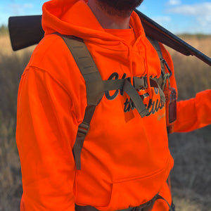 A hunter wearing a blaze orange heavyweight hoodie with 'Chasing the Flush' graphic, carrying a shotgun and wearing a hunting vest, outdoors in a field.